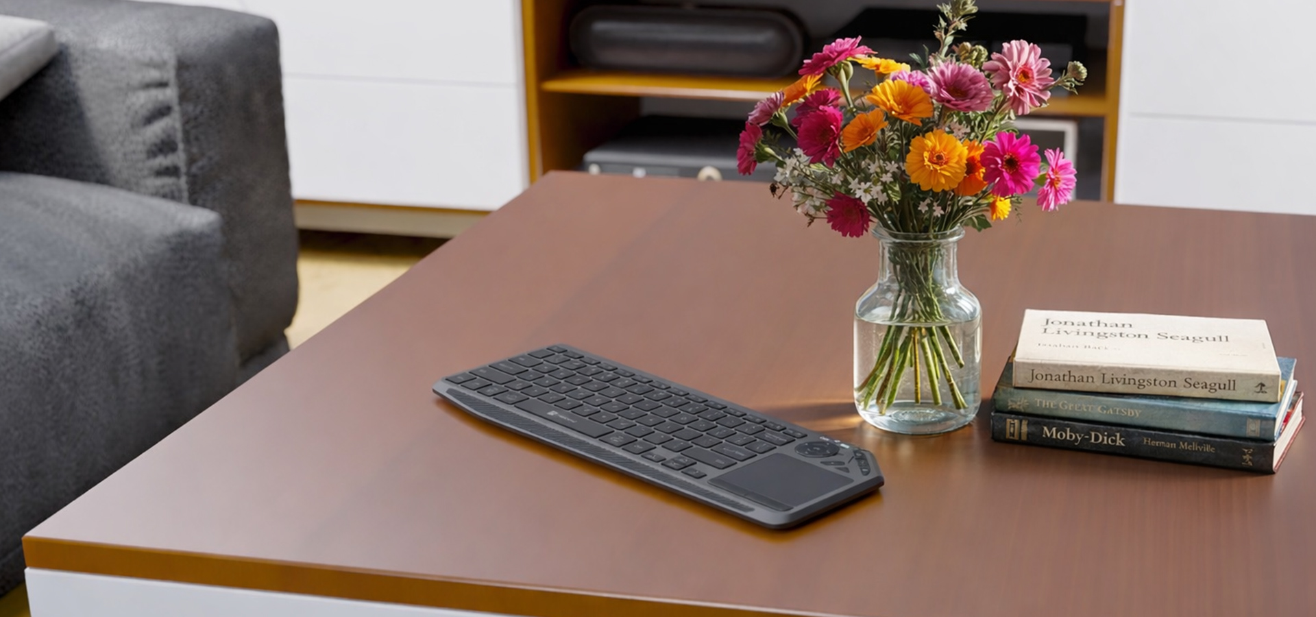 Czarna klawiatura leży na brązowym stole obok wazonu z wielokolorowymi kwiatami i książkami.