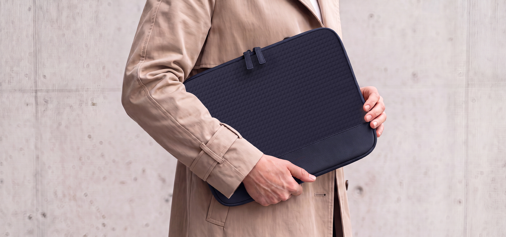 Osoba trzyma ciemną torbę na laptopa w jasnym płaszczu trenczowym.