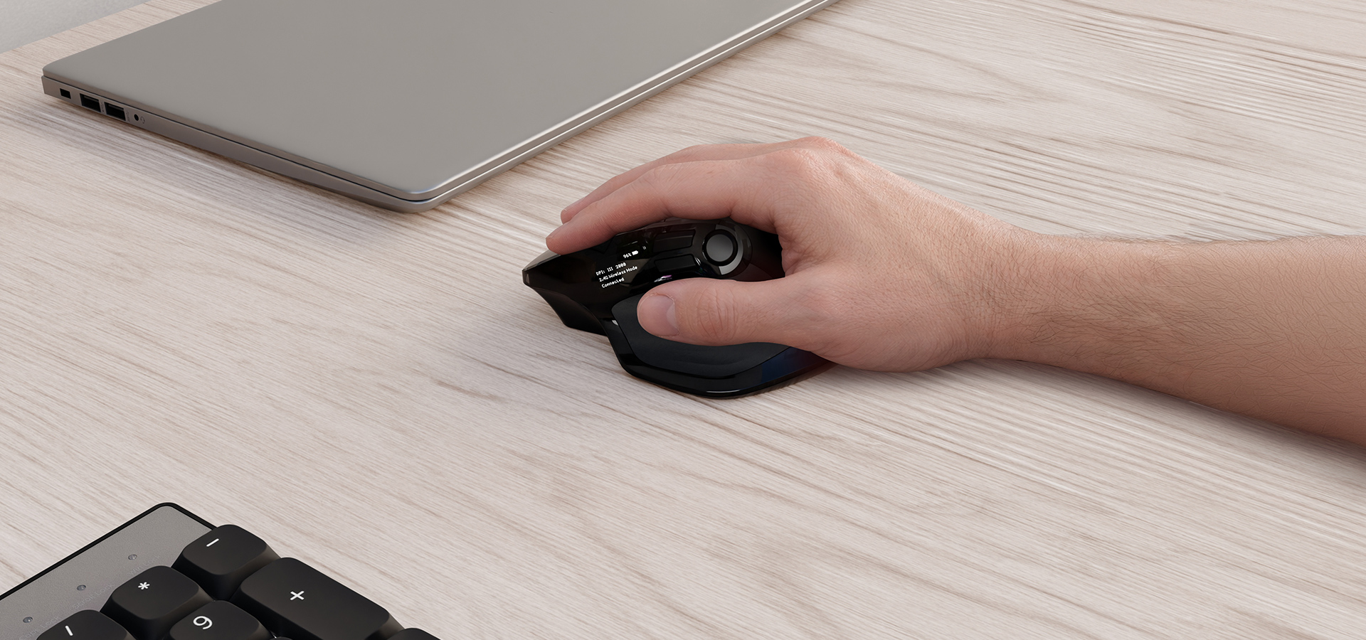 A hand using a wireless mouse on a light wooden desk beside a laptop and keyboard.