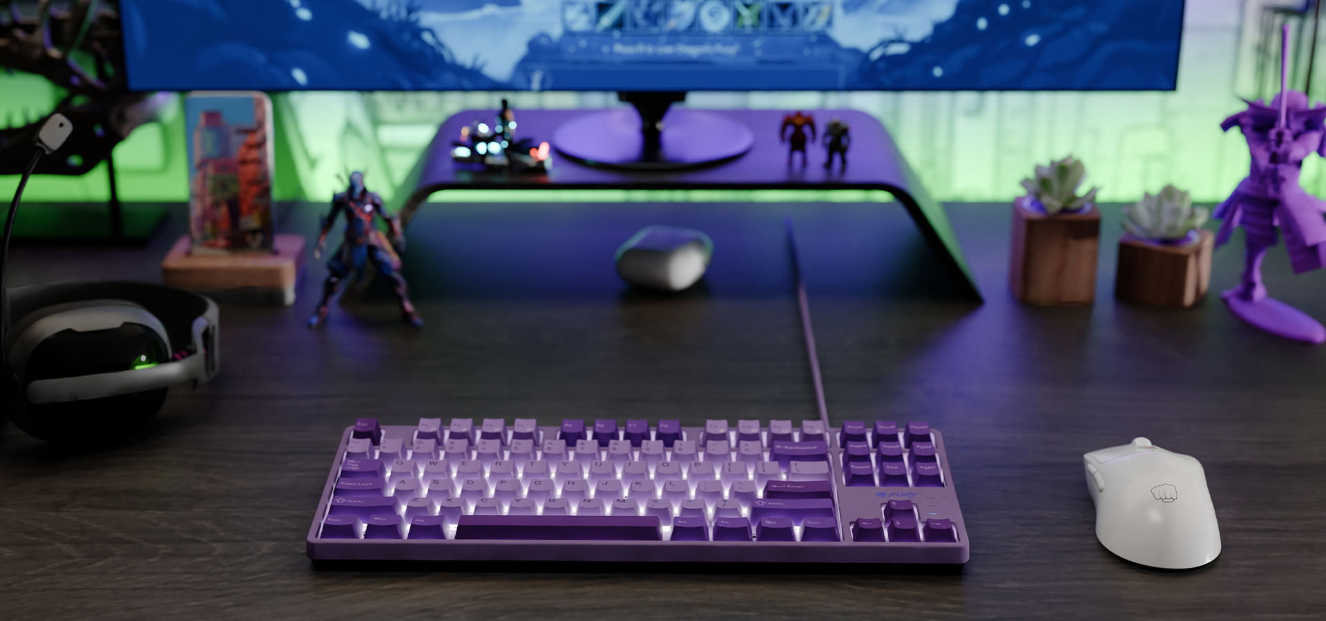 Fioletowa klawiatura z podświetlonymi klawiszami obok białej myszy na ciemnym biurku.