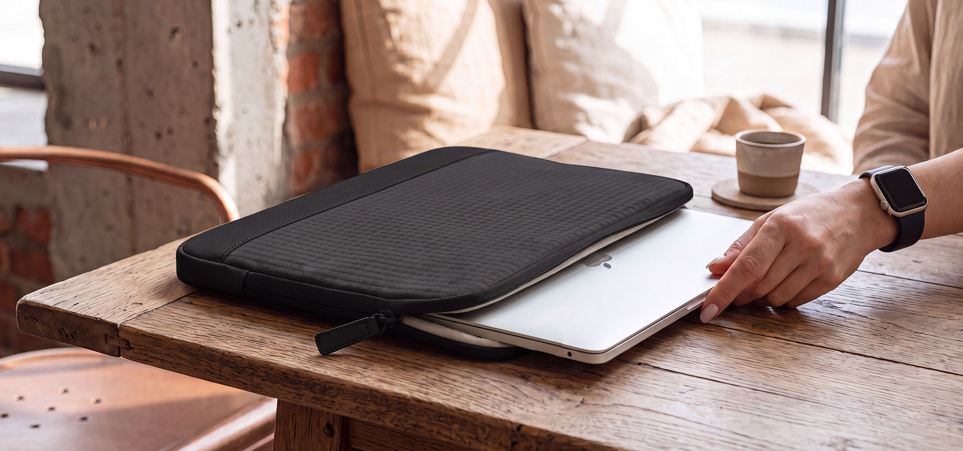 Osoba wkłada laptopa do czarnej torby na drewnianym stole obok filiżanki.
