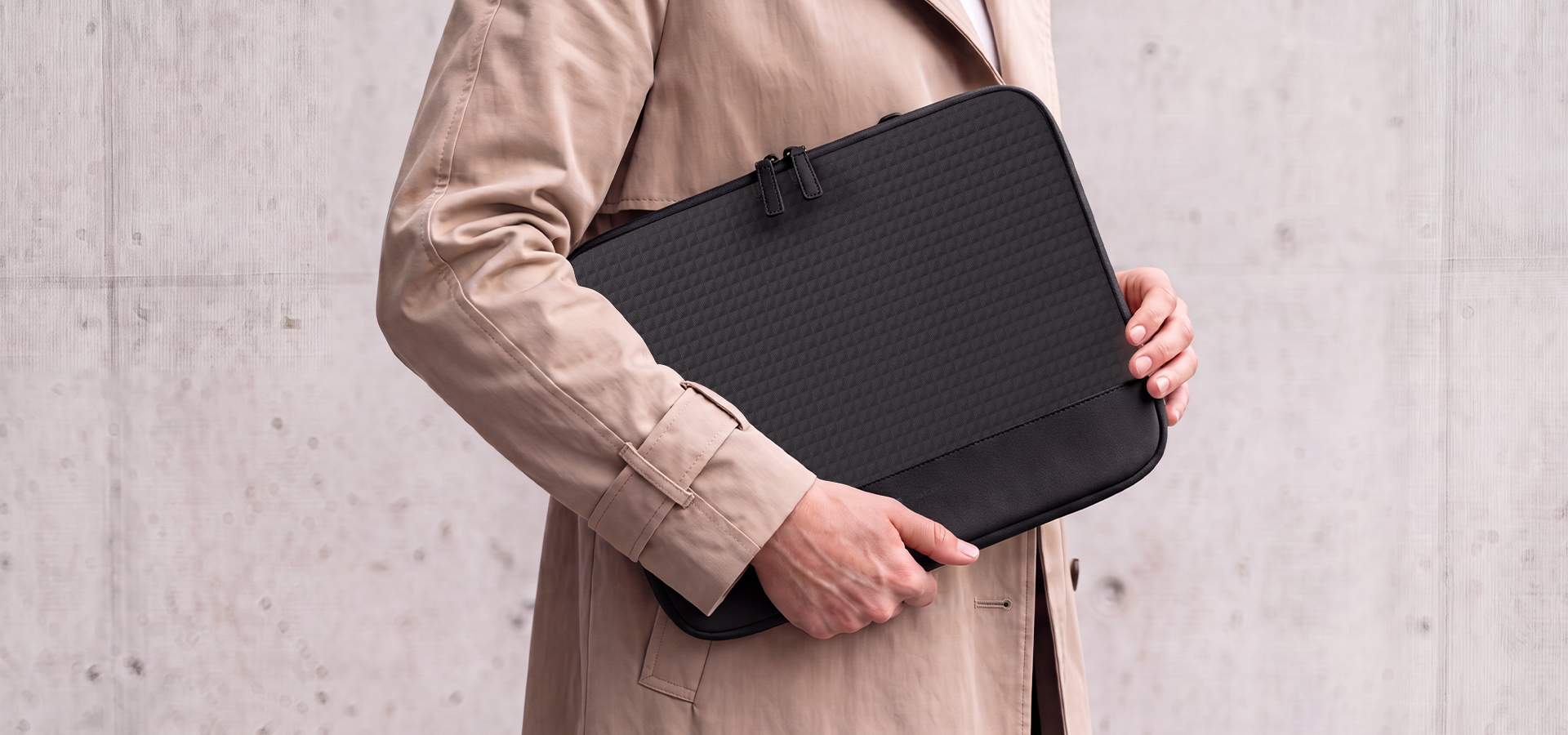 Osoba trzyma czarną torbę na laptopa przy teksturowanej betonowej ścianie.