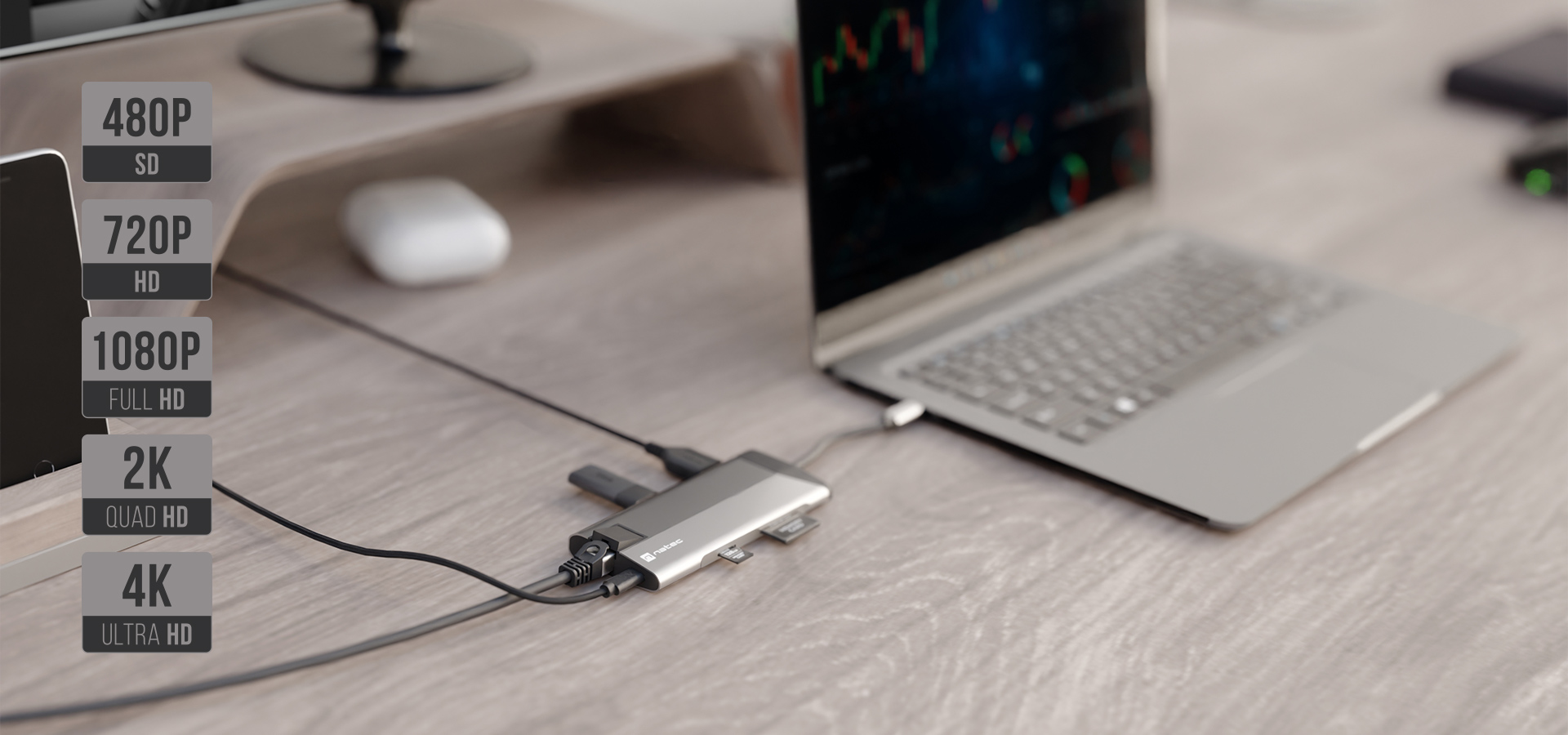 Laptop podłączony do huba wieloportowego na drewnianym biurku z monitorem w tle.