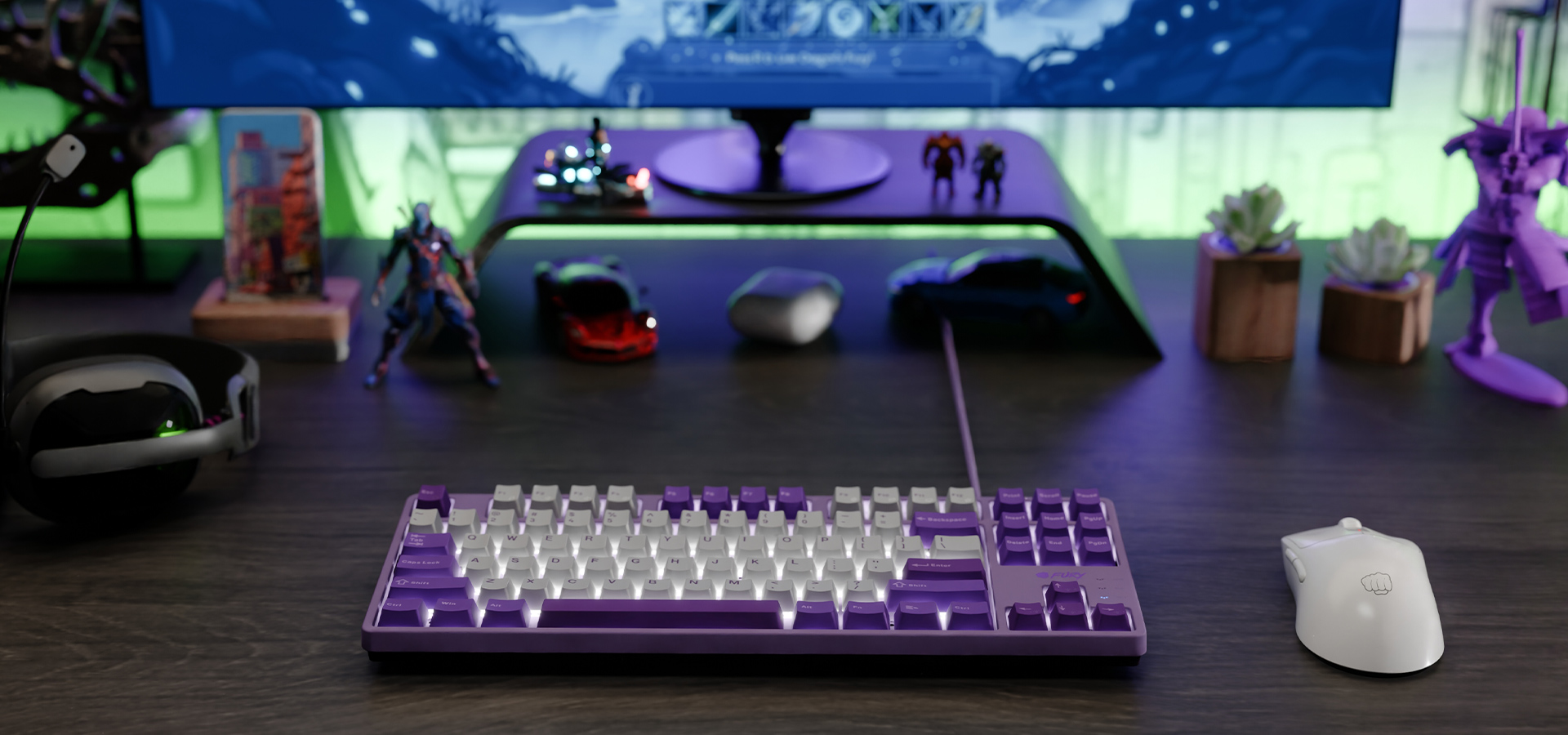 Fioletowa klawiatura i biała myszka na biurku z figurkami i monitorem w tle.